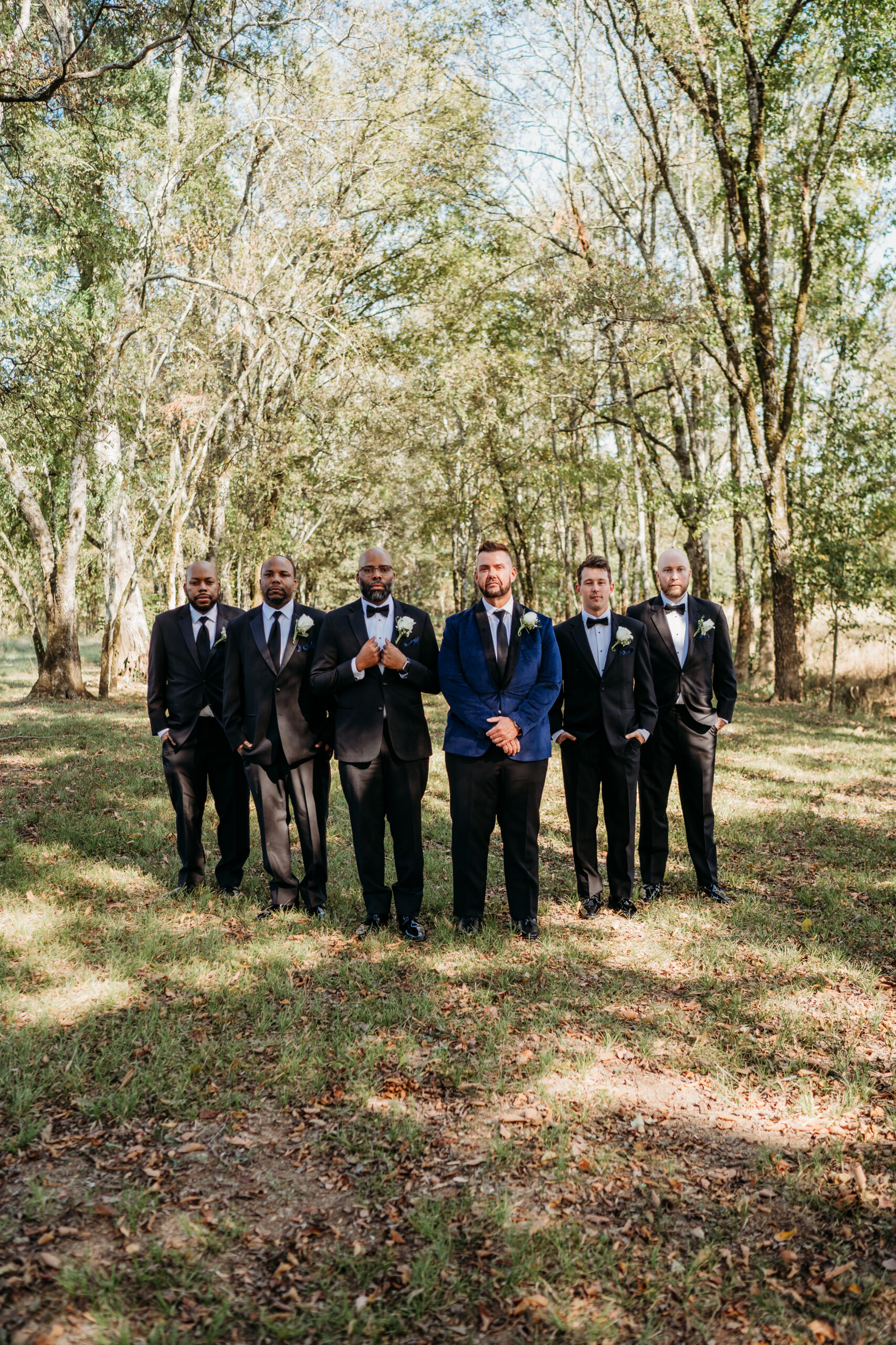 Levi & Billy with four groomsmen in black tuxedos pose among leaf-strewn Covey Creek trees