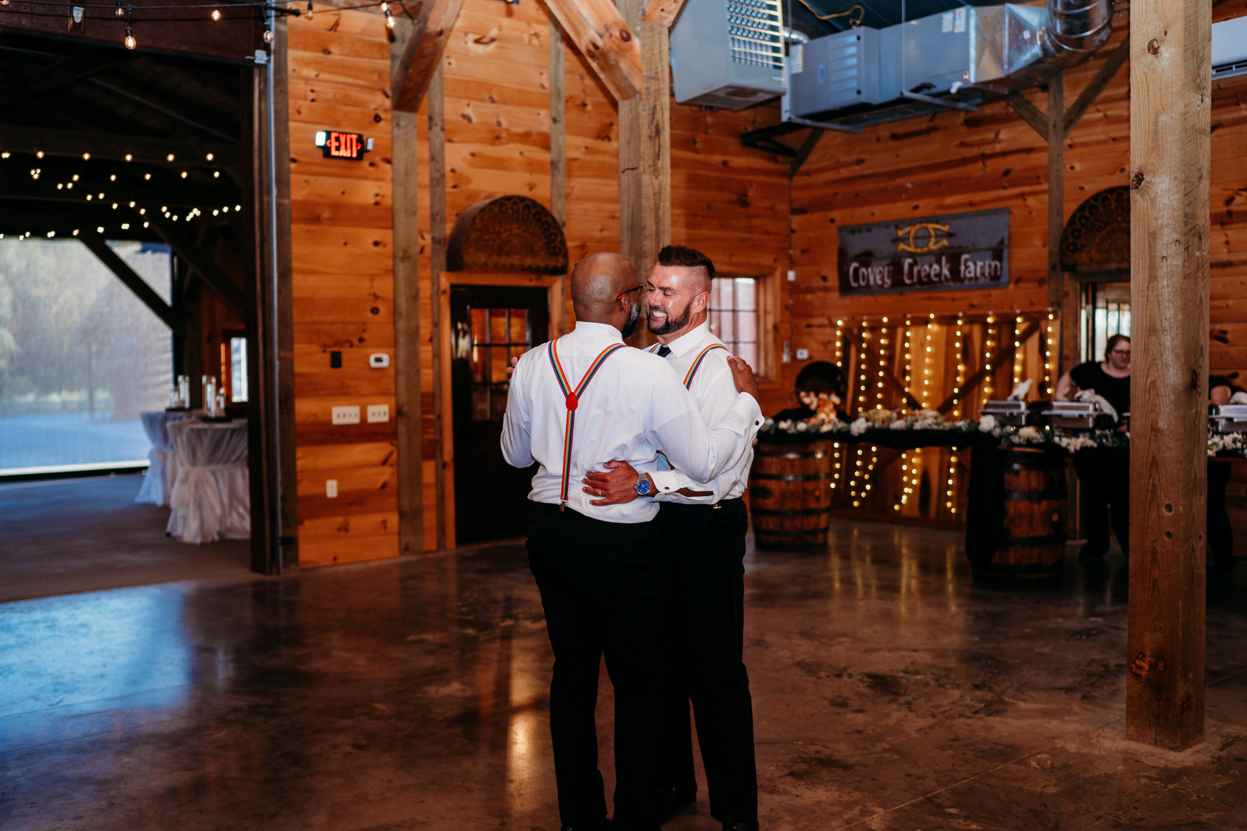 Levi & Billy embrace dancing in rainbow suspenders beneath Covey Creek Farm wall sign indoors