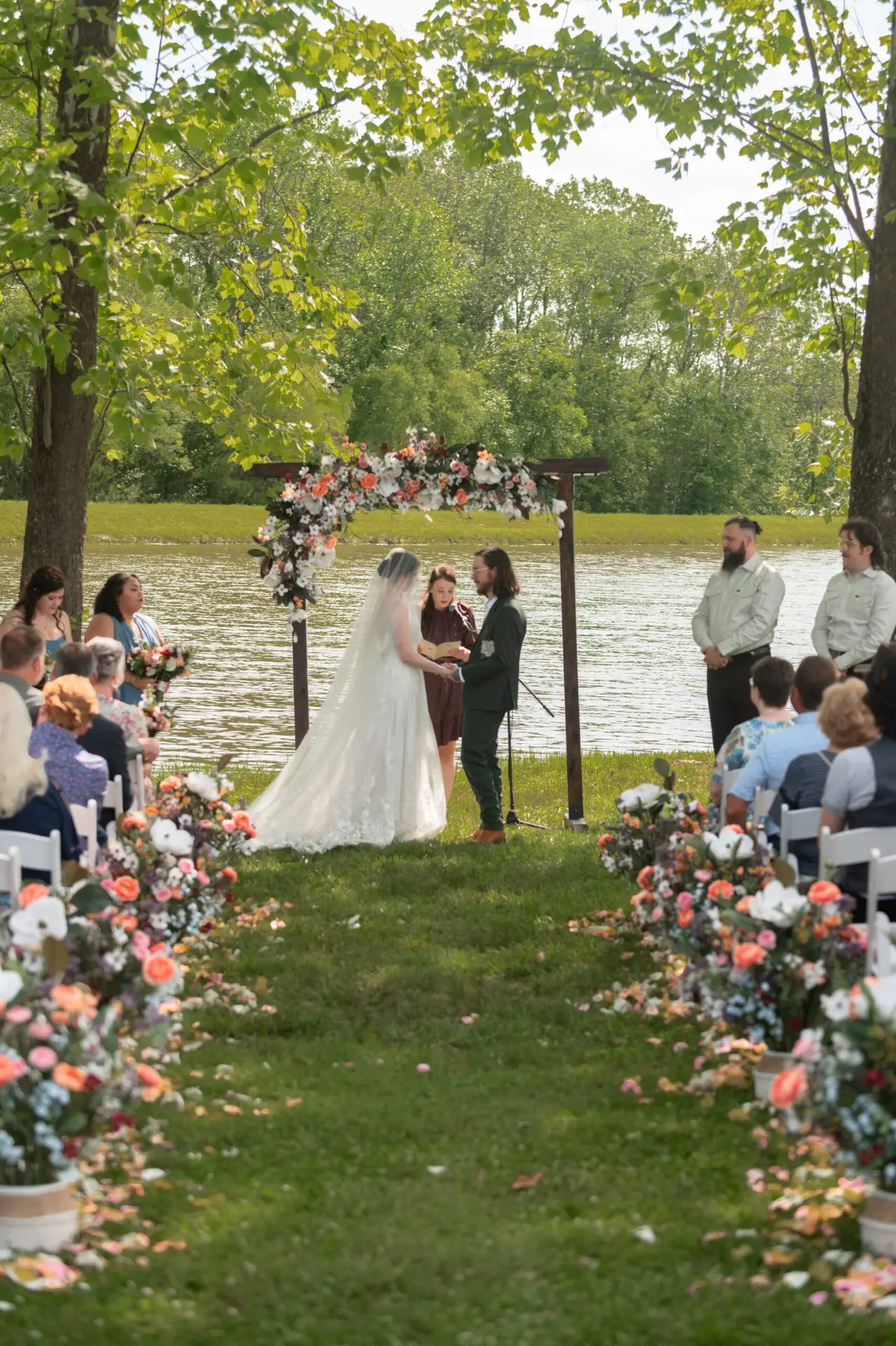 Pondside ceremony for Rachael & Ben under a floral wood arbor with aisle florals at Covey Creek.