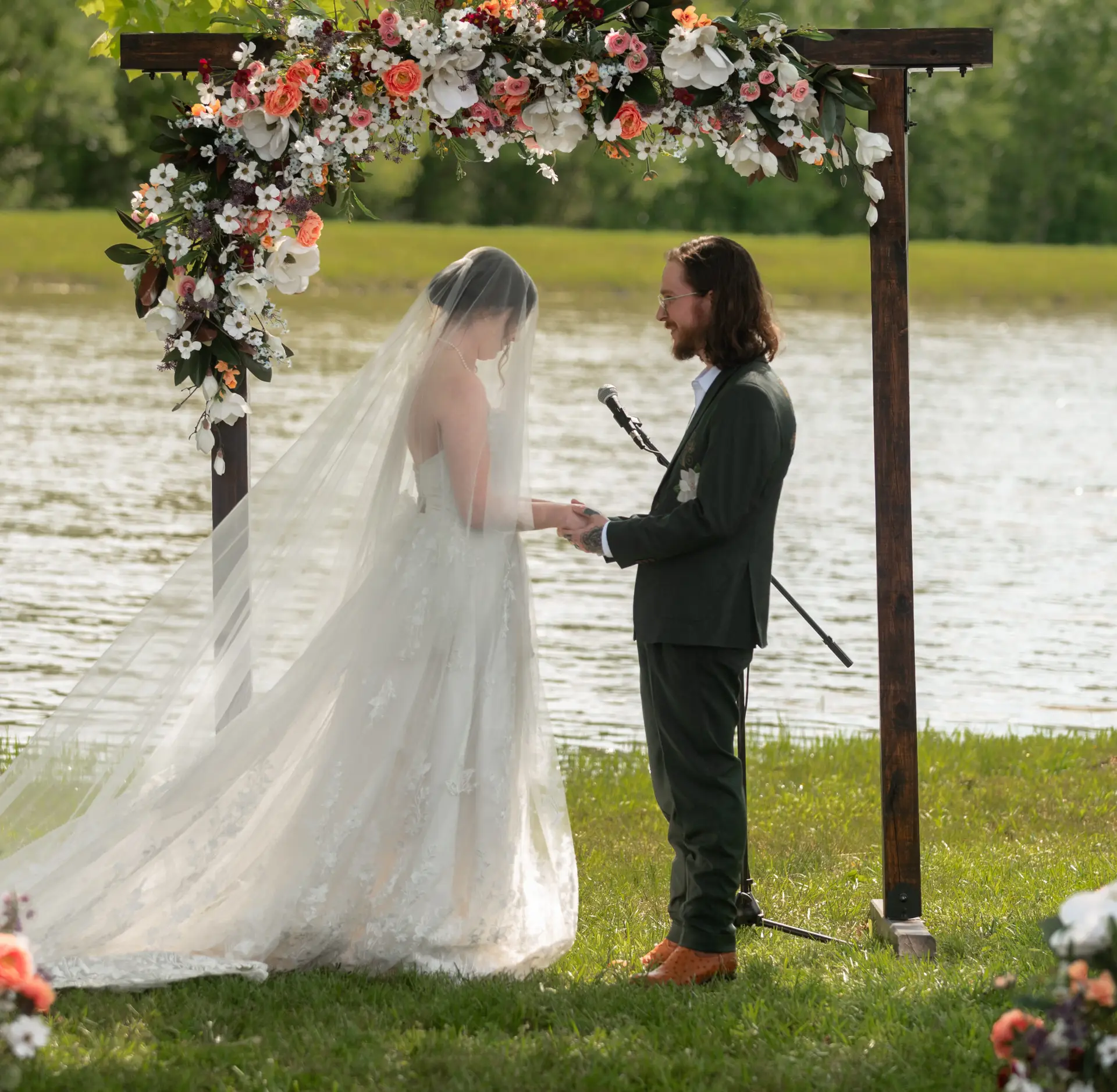 Rachael & Ben hold hands under a floral wood arbor by the pond during their ceremony at Covey Creek.
