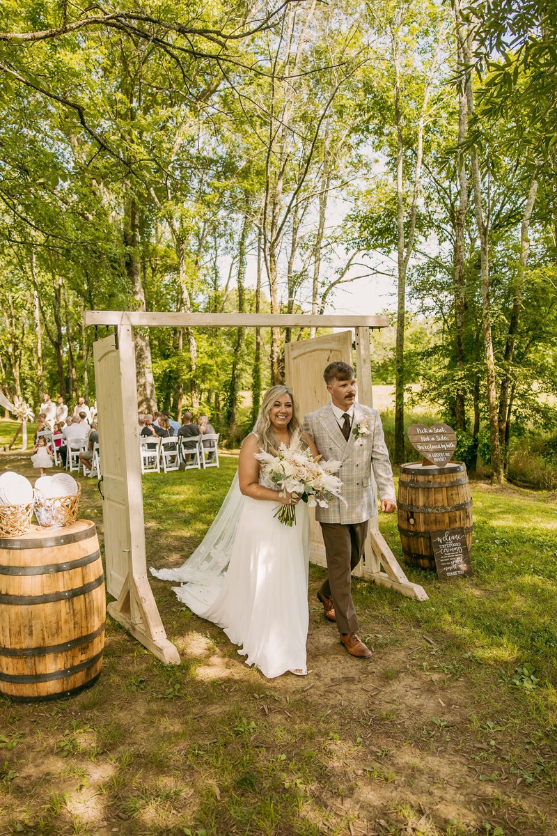 Alexis & Logan exit through open white doors past whiskey barrels after the ceremony at Covey Creek