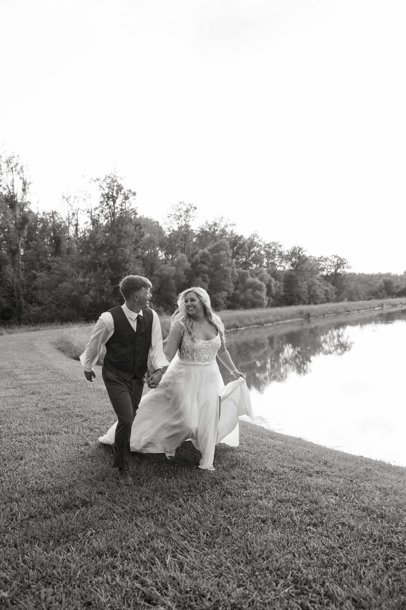 Alexis & Logan run hand in hand along the grassy pond bank in a black-and-white portrait at Covey Creek