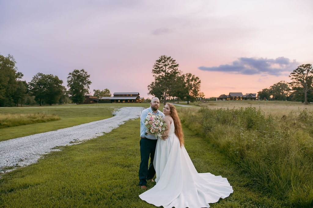 Amanda & Stephen stand in a Covey Creek field at twilight, barns glowing along the gravel road behind