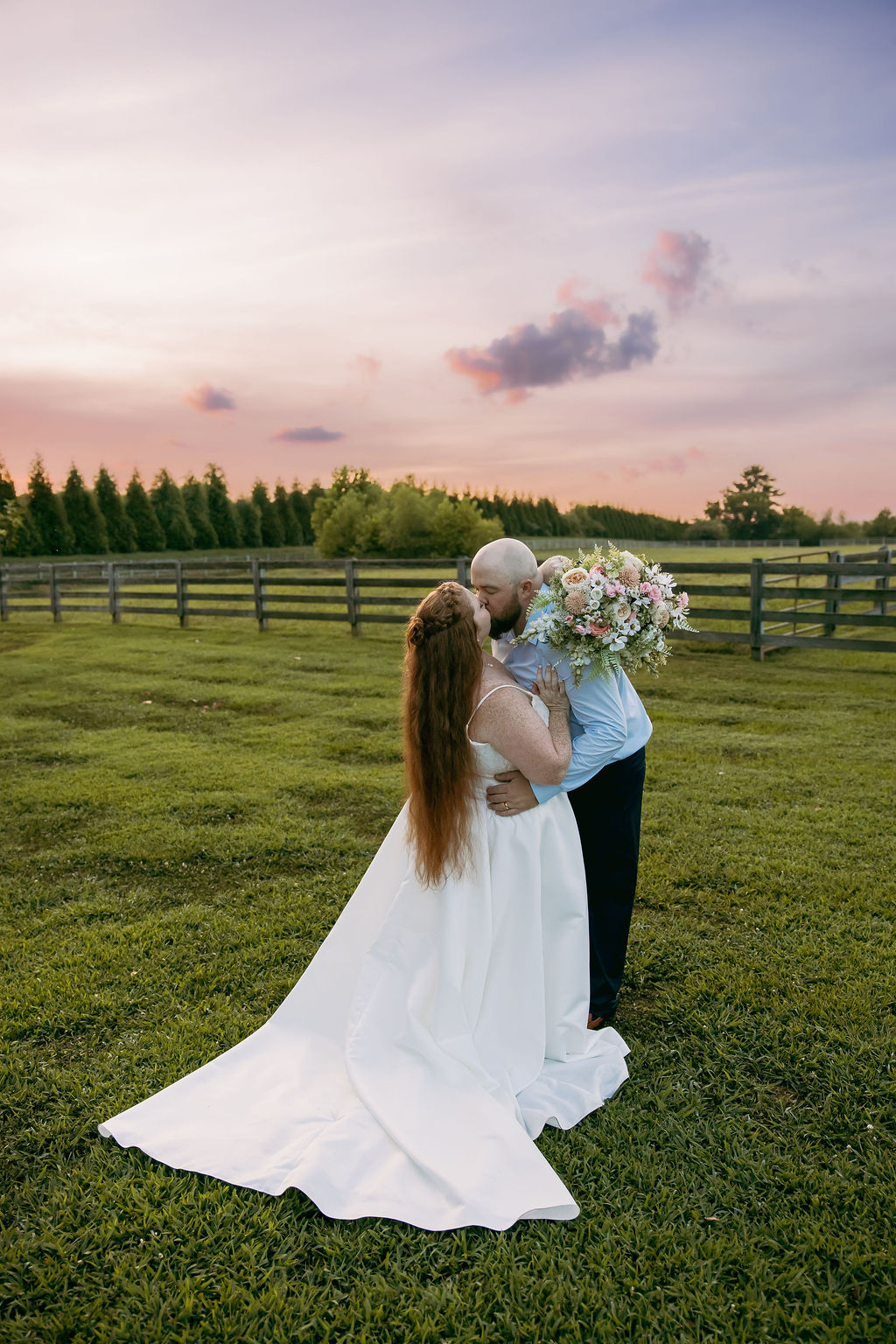 Amanda & Stephen kiss in a Covey Creek field at sunset beside a wooden fence and evergreen treeline
