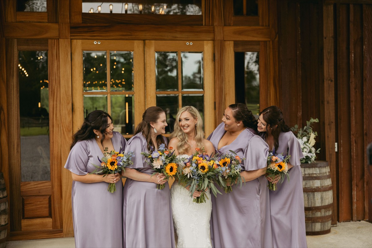 Brianna & Michael's bridal party: she laughs with lavender bridesmaids and sunflower bouquets at Covey Creek's doors.