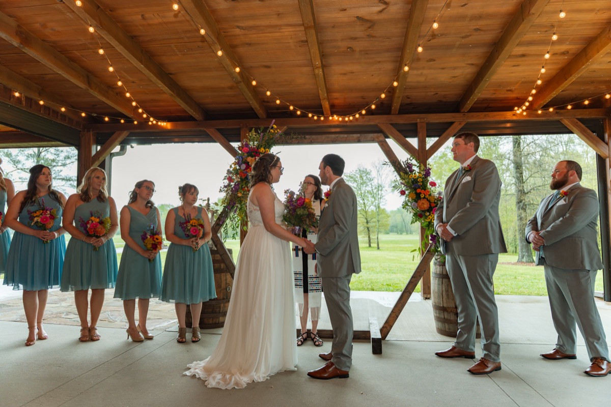 Caitlin & Daniel hold hands under string lights at the Pond pavilion with teal bridesmaids at Covey Creek