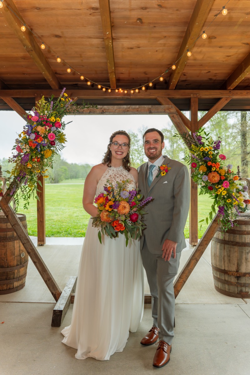 Caitlin & Daniel pose under a floral hex arch on whiskey barrels at the Pond pavilion at Covey Creek