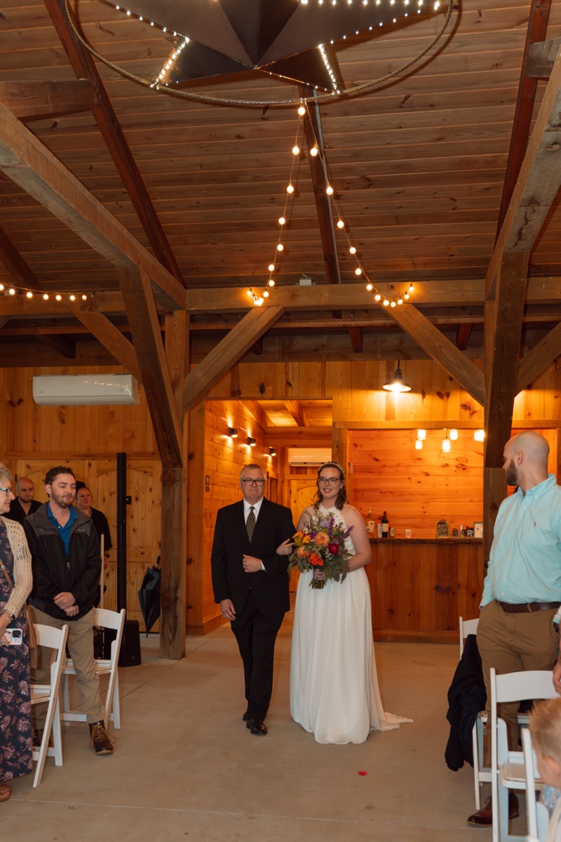 Caitlin & Daniel: bride walks the barn aisle arm-in-arm with her father under string lights at Covey Creek