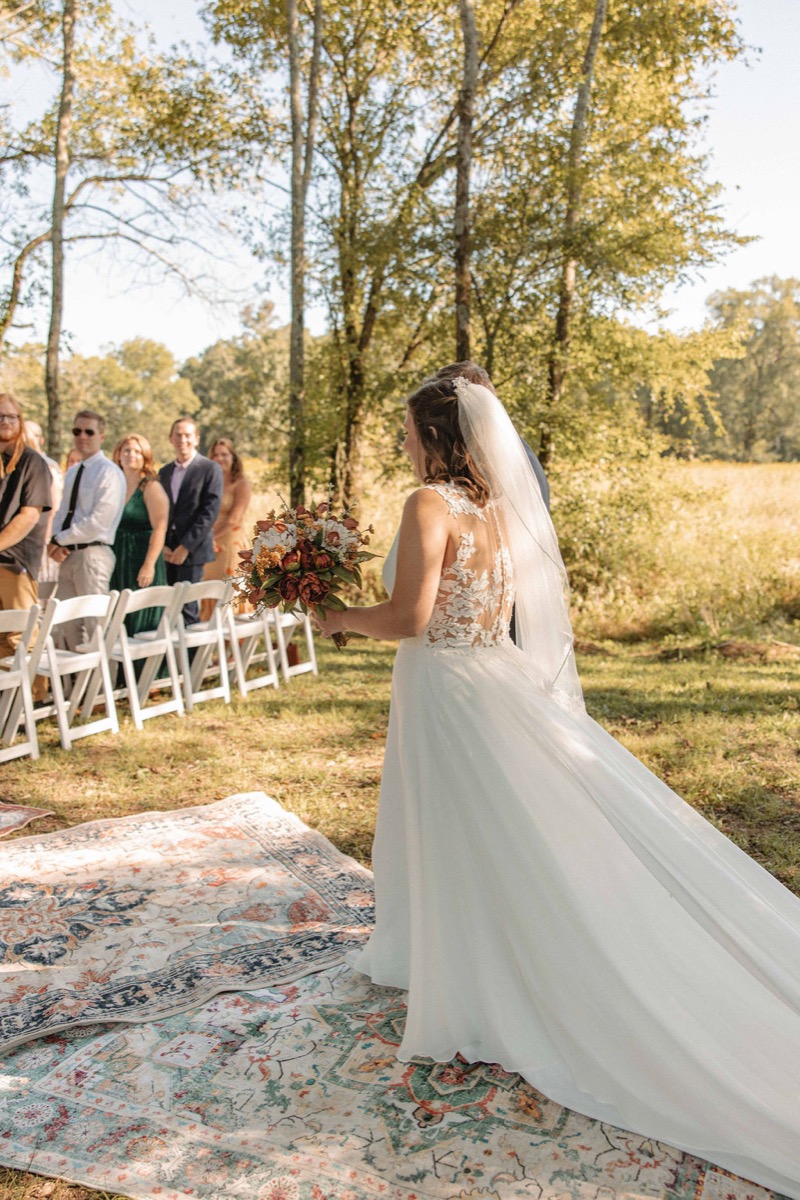 Claire & Jacob's processional as Claire walks a patterned-rug aisle under trees at Covey Creek