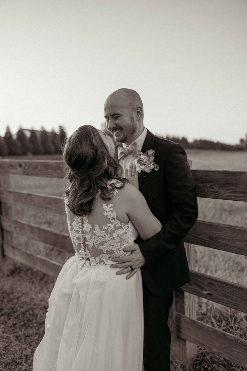 Claire & Jacob embrace by a wooden fence, her lace-back gown toward the camera at Covey Creek