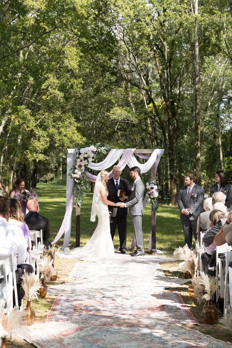 Courtney & Brett exchange vows under a fabric-draped wooden arbor along the rug aisle at Covey Creek