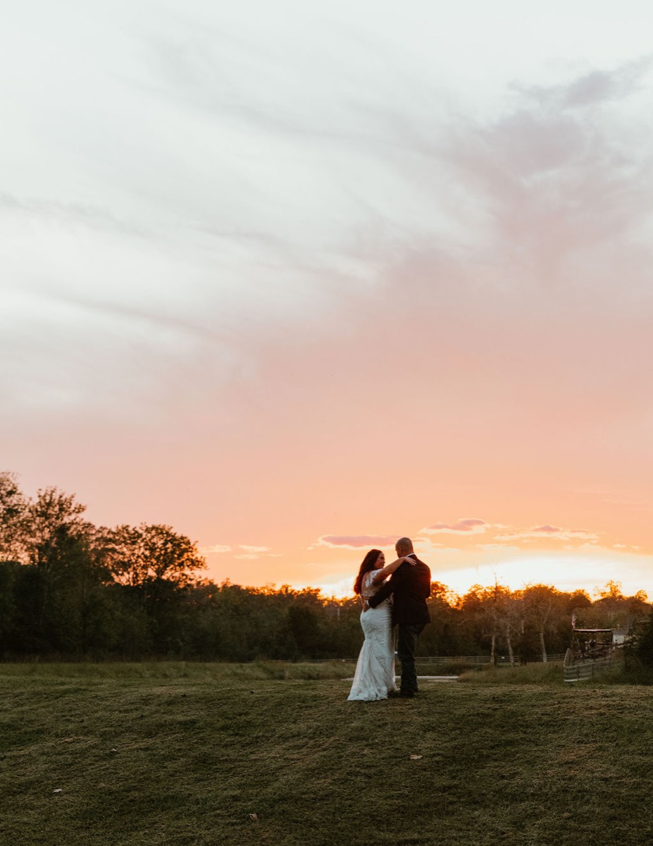 Elizabeth & Ashton embrace on a grassy hill at Covey Creek beneath a glowing peach-and-orange sunset sky