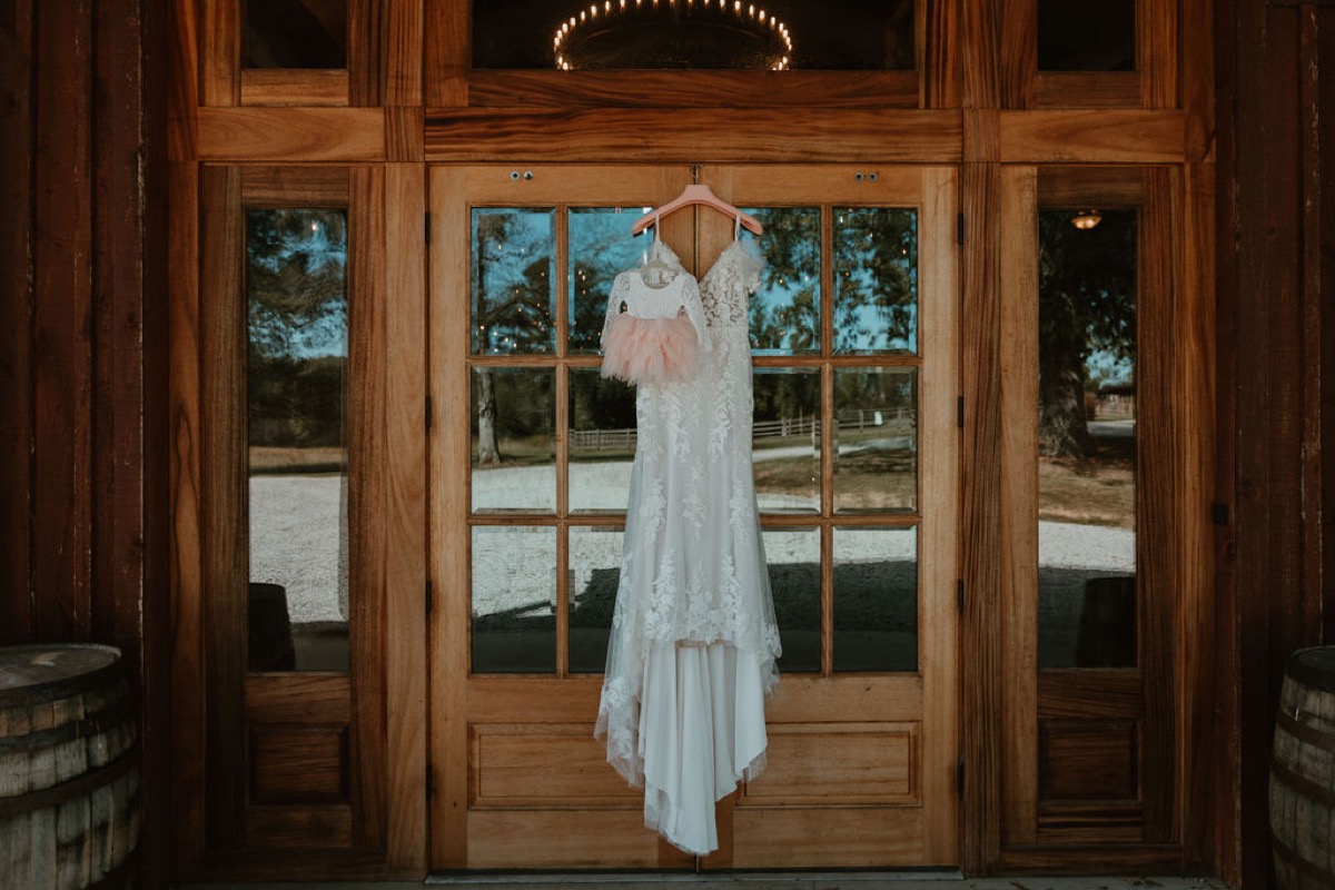 Elizabeth & Ashton’s lace gown and pink flower-girl dress hang on Covey Creek’s glass-paneled wooden barn doors