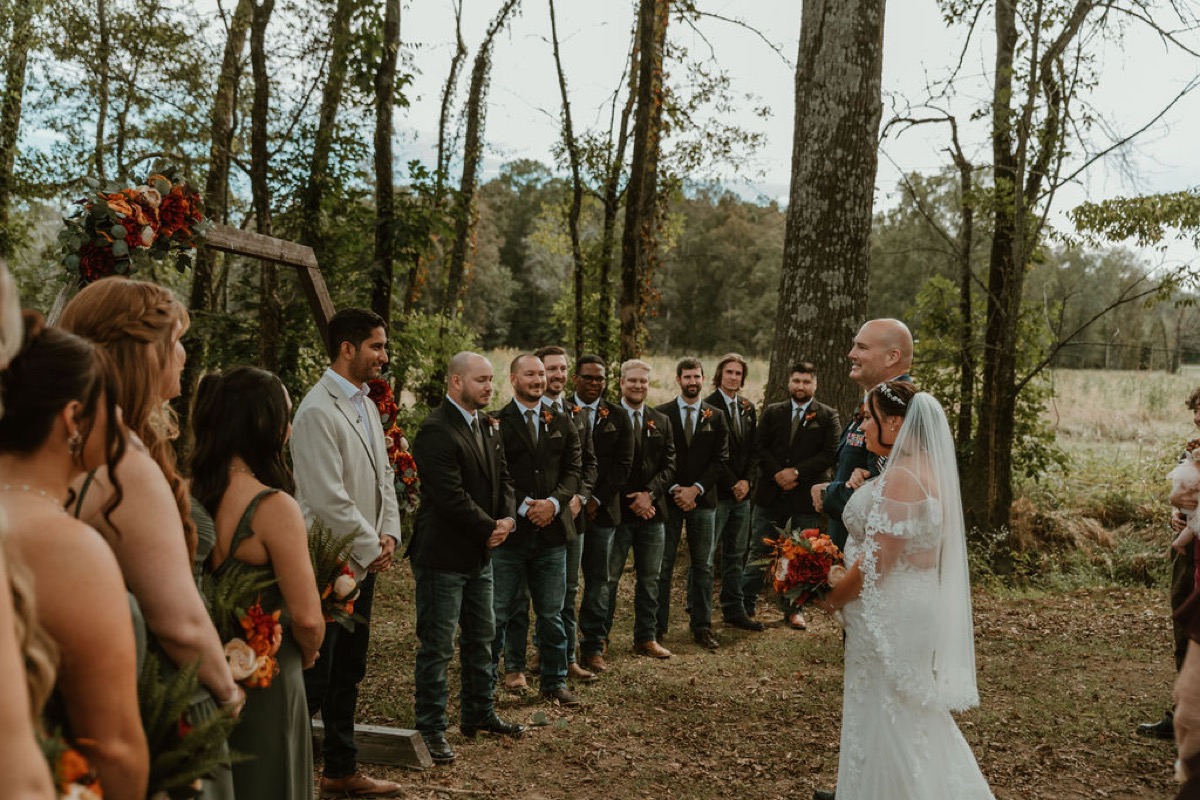 Elizabeth & Ashton stand with their wedding party under a flowered hex arbor in Covey Creek’s wooded clearing
