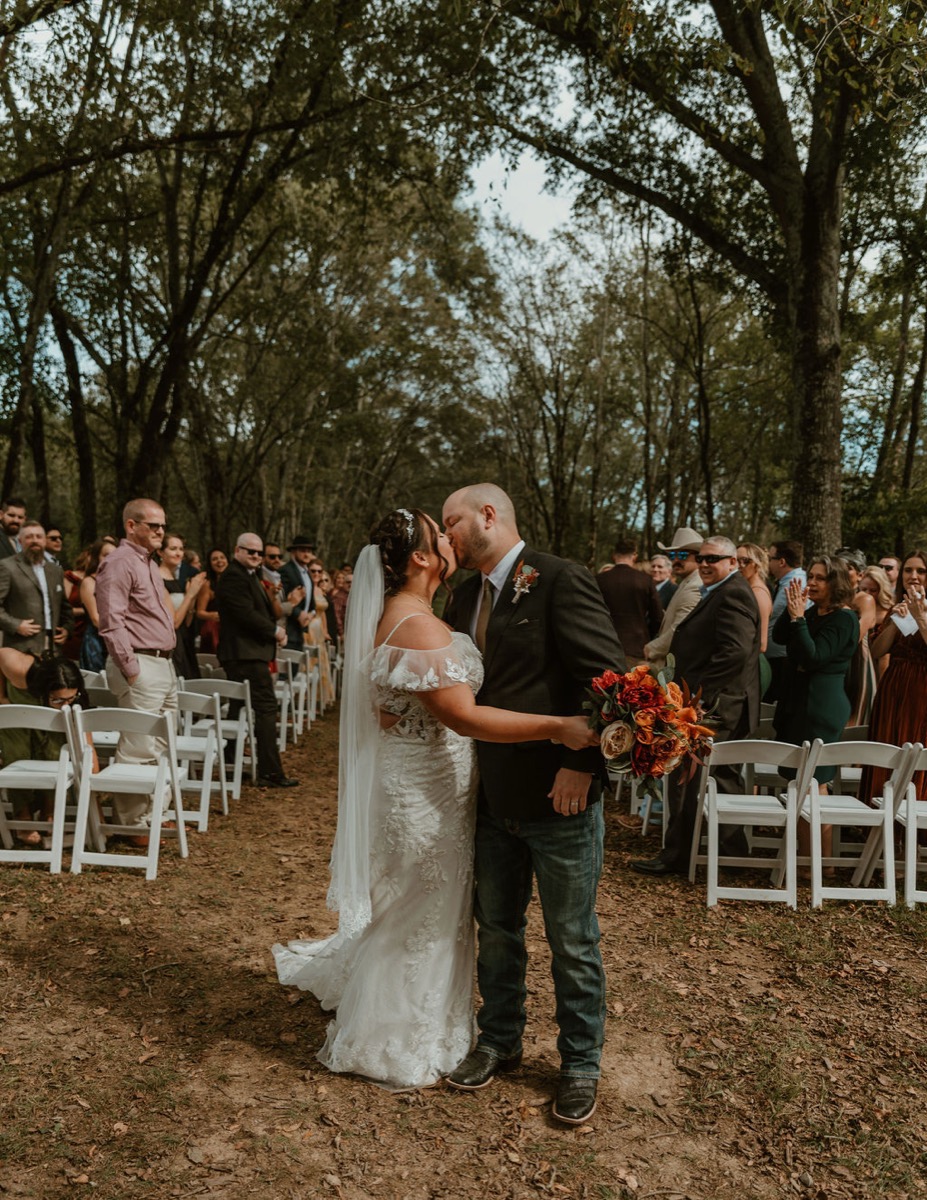 Elizabeth & Ashton kiss on the wooded aisle at Covey Creek, autumn bouquet and white chairs framing them