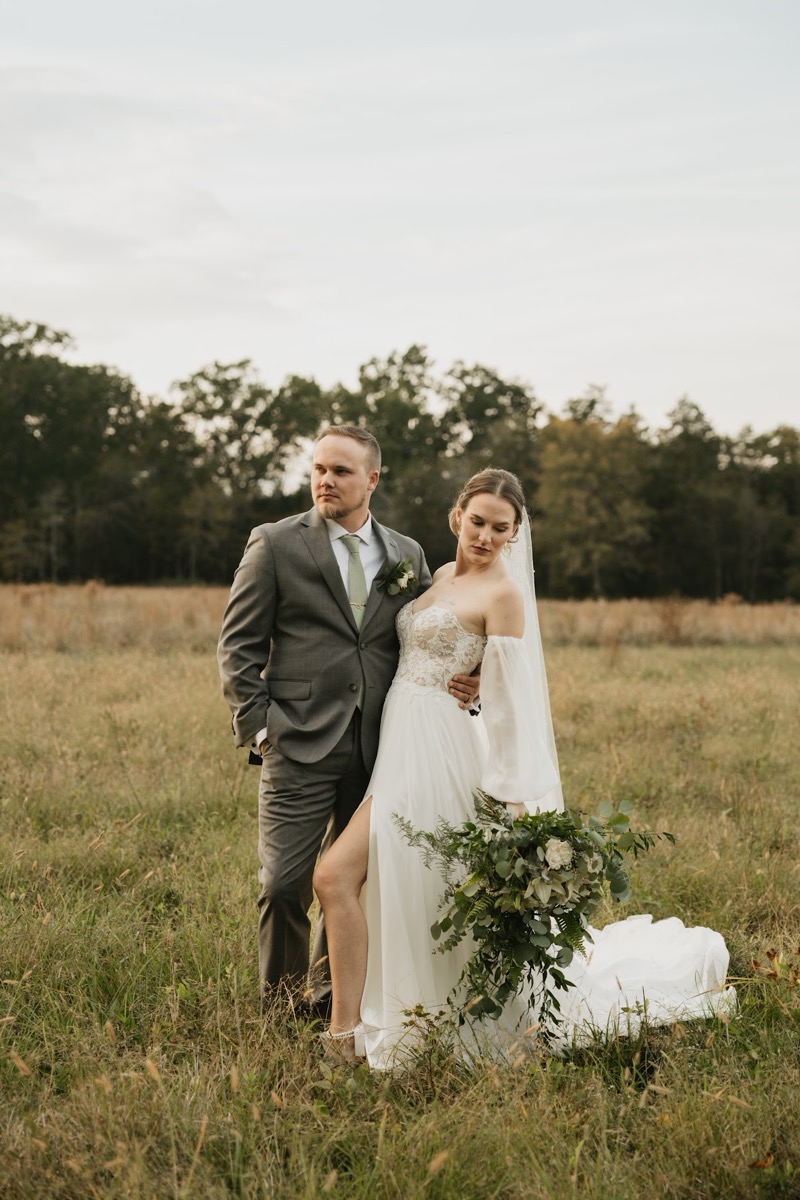 Emily & Bryan pose in tall golden grass, she with a cascading bouquet and puff sleeves, he in a grey suit at Covey Creek.