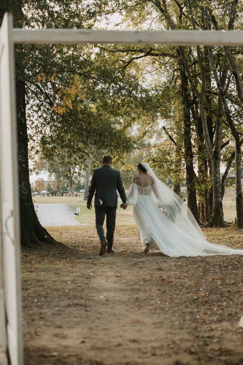 Emily & Bryan walk hand-in-hand down the Tunnel of Love path, her long veil trailing past a white doorway at Covey Creek.