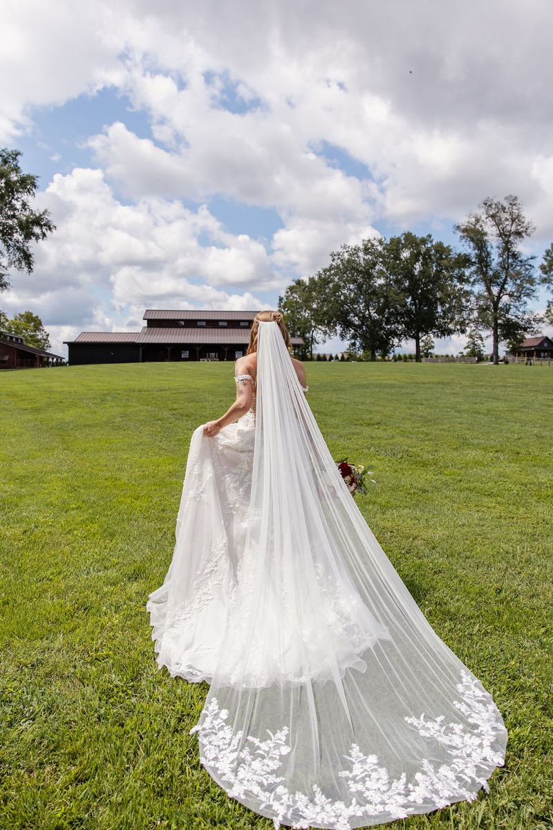 Erica & Randall’s bride walks toward the dark barn, long lace veil trailing across the Covey Creek lawn