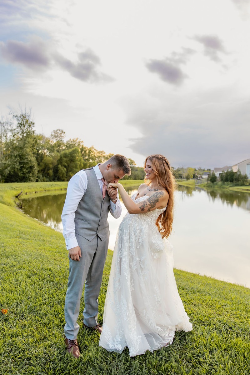 Erica & Randall pose beside Covey Creek Pond as he kisses her hand on the grassy bank under cloudy skies