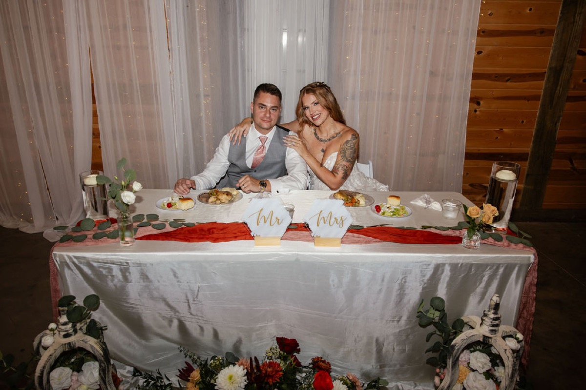 Erica & Randall sit at their sweetheart table with Mr & Mrs signs and fairy-lit curtains at Covey Creek