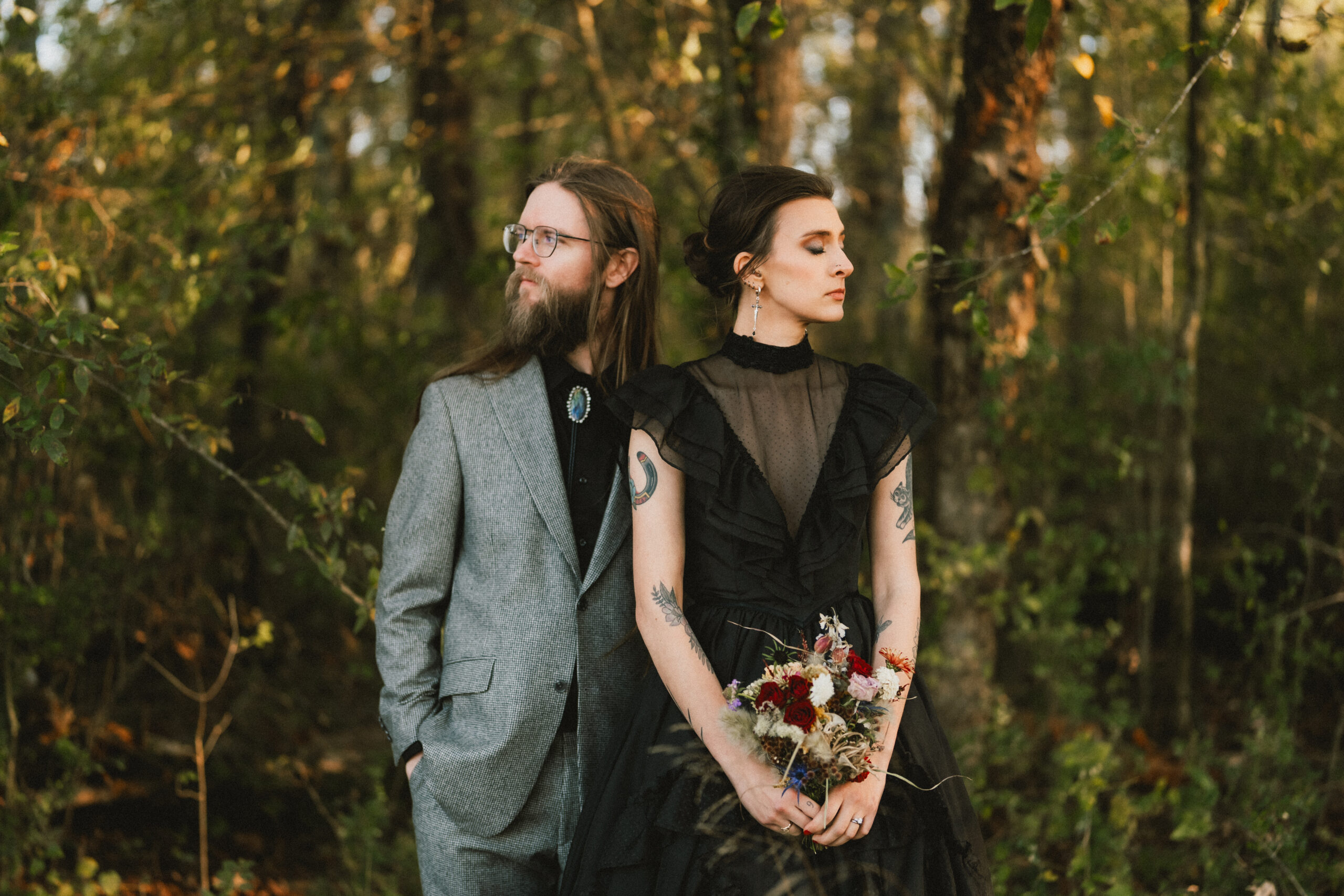 Erin & Baxter stand back-to-back in sunlit woods, her black gown and dried bouquet at Covey Creek