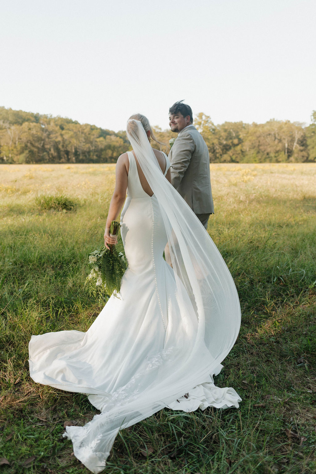 Hailey & Joseph walk through a grassy field, her long veil trailing behind, at Covey Creek