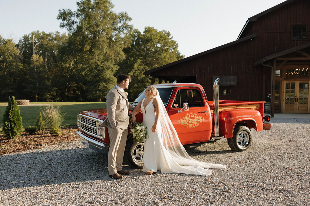 Hailey & Joseph pose by a vintage red pickup with the Covey Creek barn glowing behind them at golden hour