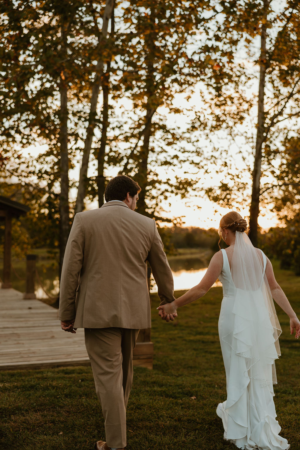 JJ & Will walk hand in hand toward the Pond at sunset, gown train and tan suit backlit at Covey Creek