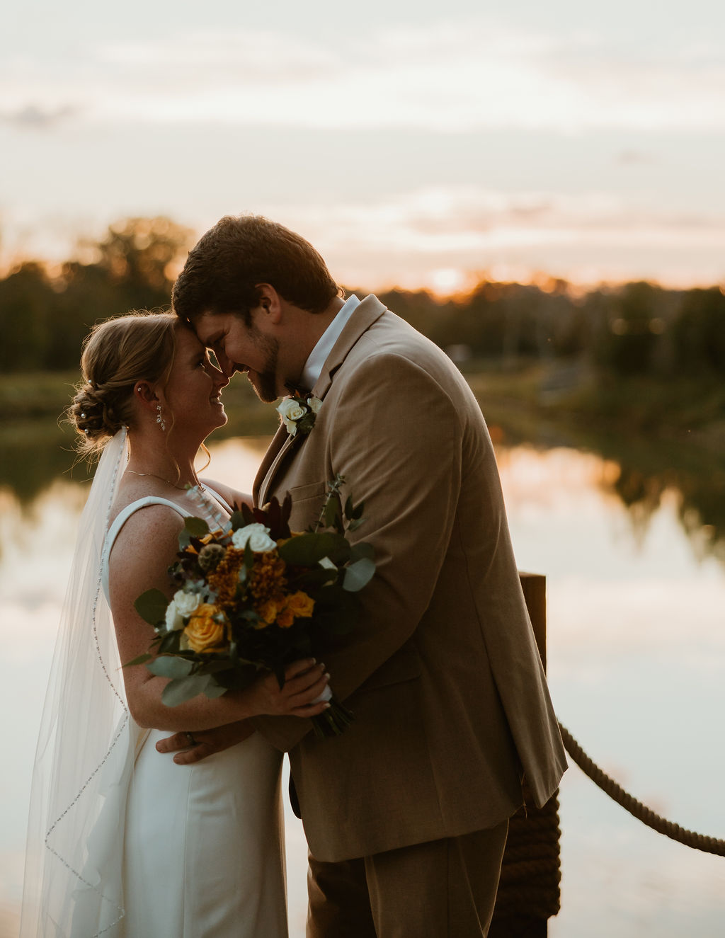 JJ & Will touch foreheads on the Pond dock at golden hour, bouquet and tan suit at Covey Creek