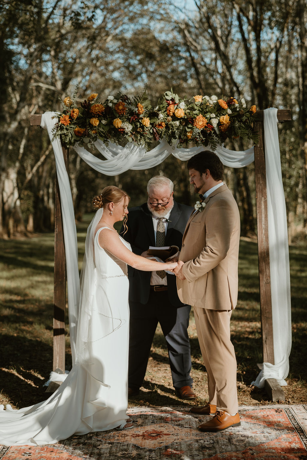 JJ & Will hold hands under a floral-draped arbor as the officiant reads vows at Covey Creek