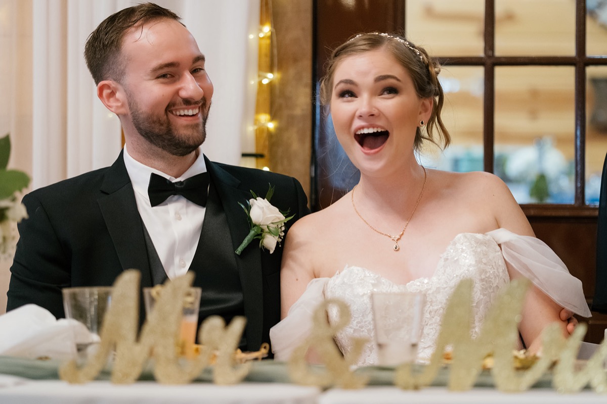Katelyn & Josh laugh at their Covey Creek head table beside a glitter Mr & Mrs sign and string lights