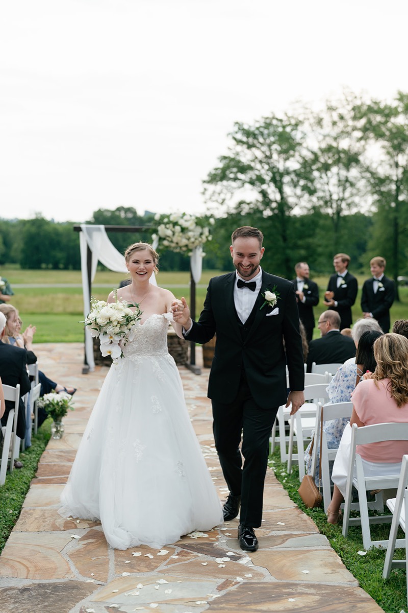 Katelyn & Josh recess down Covey Creek’s Patio aisle past guests, white petals on the flagstone path