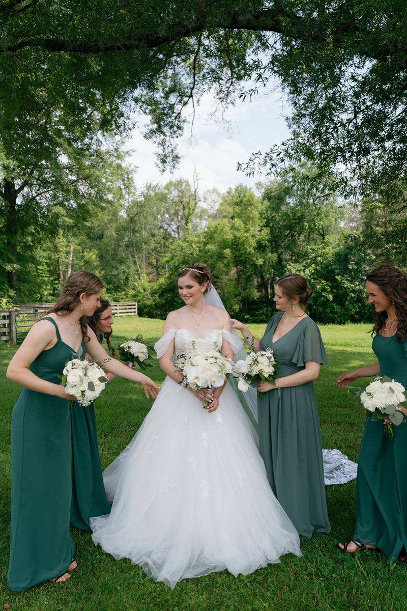 Katelyn & Josh bridesmaids fluff the bride’s veil and gown under a tree on Covey Creek’s lawn
