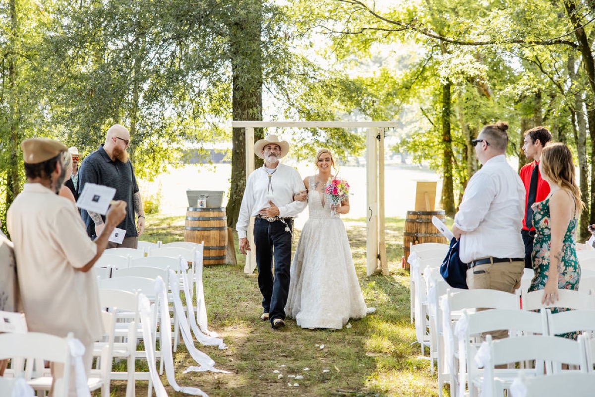 Kolbe walks the grassy aisle with her father past wooden doors under trees at Covey Creek, Kolbe & Nathan