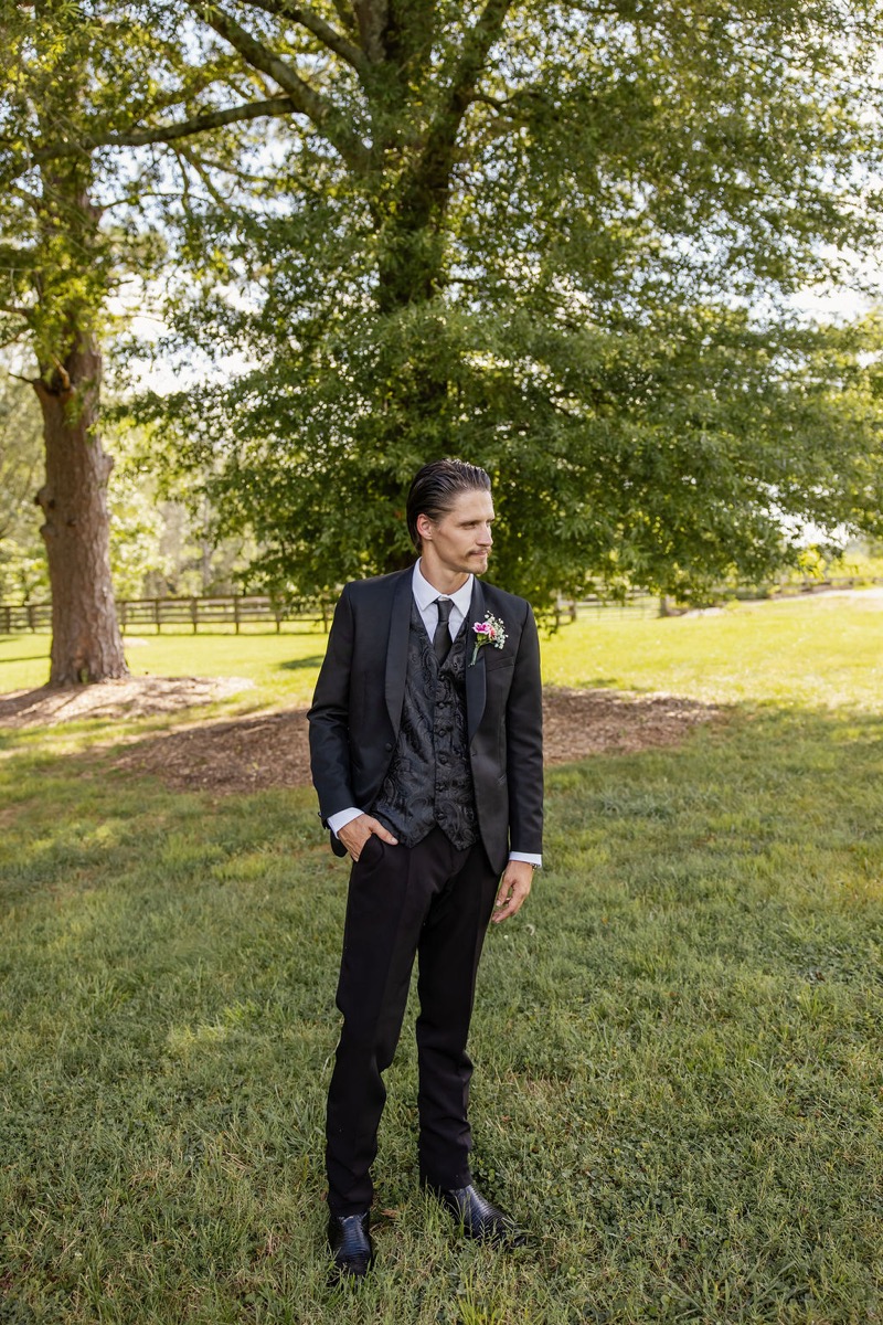 Nathan in a black suit and brocade vest stands on the lawn at Covey Creek for Kolbe & Nathan's wedding