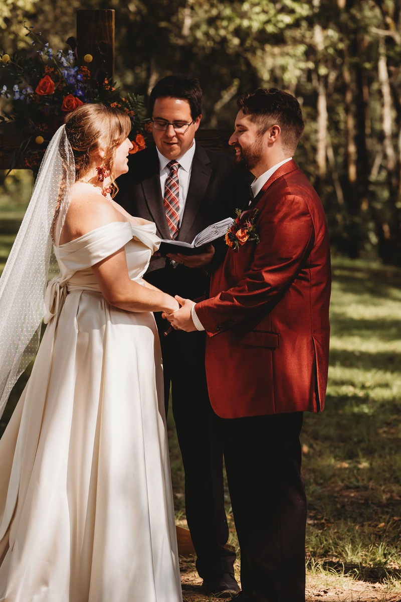 Lauren & Alex hold hands at the altar before a floral wooden cross during vows at Covey Creek