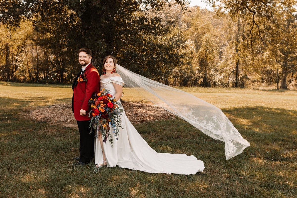 Lauren & Alex stand back-to-back in a grassy field as her long dotted veil blows at Covey Creek