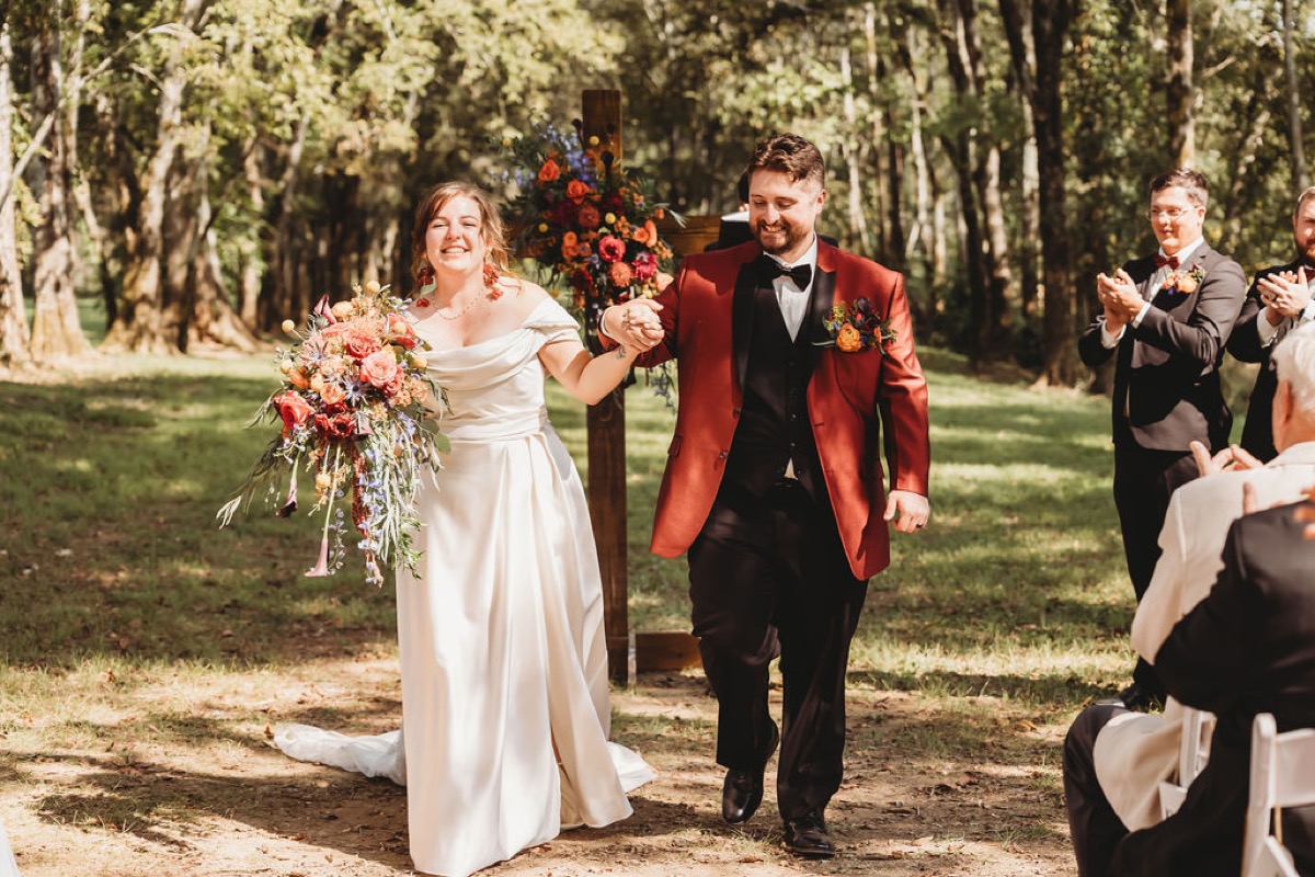 Lauren & Alex smile hand-in-hand down the grassy aisle after vows as guests clap at Covey Creek