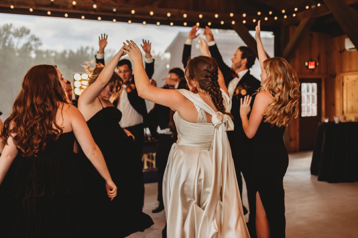 Lauren of Lauren & Alex raises her arms dancing in a circle of guests in the barn at Covey Creek