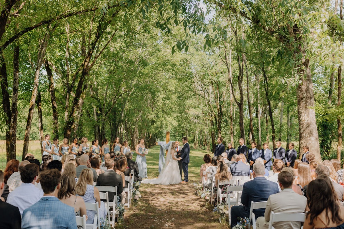 Leah & Gordon stand at the altar under Covey Creek’s arched tree canopy as guests face the aisle
