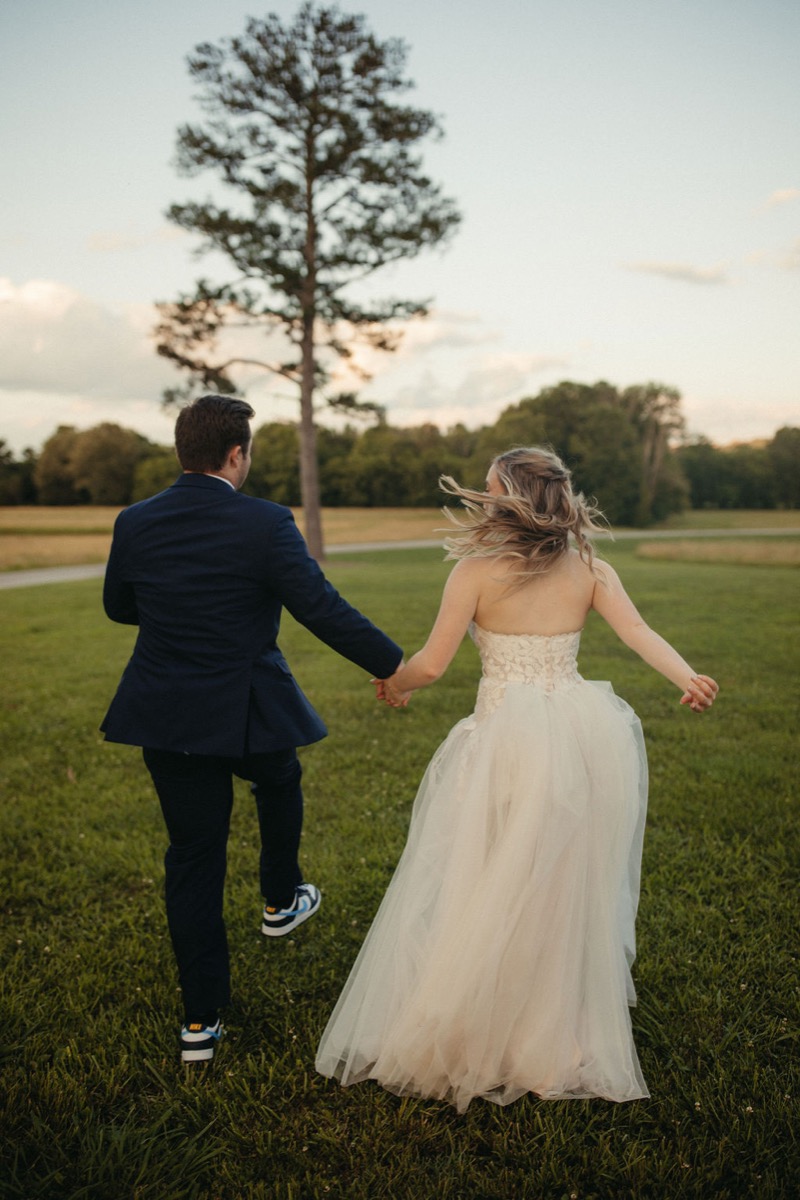 Leah & Gordon hold hands crossing a Covey Creek lawn toward a tall pine in soft golden-hour light