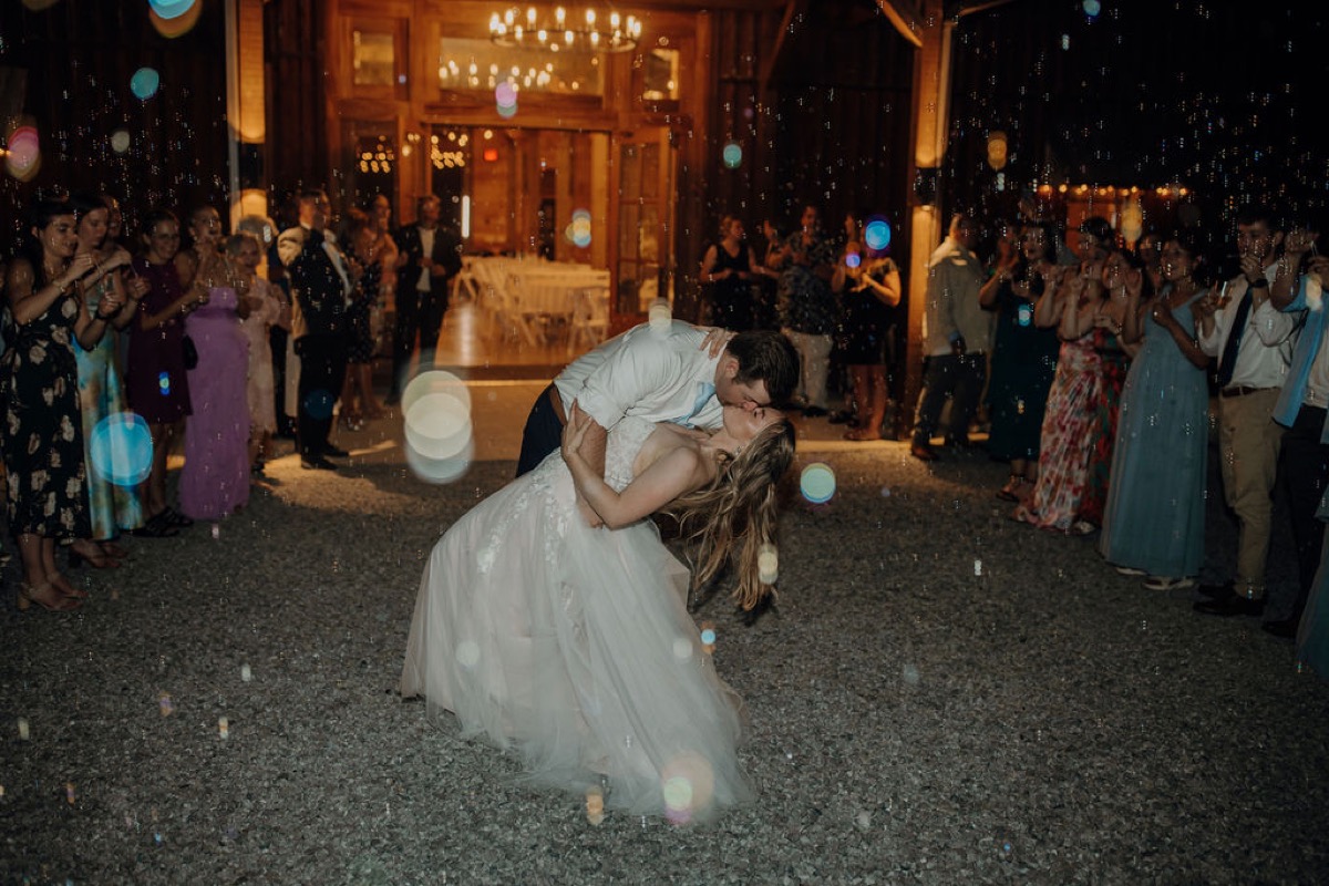 Gordon dips Leah for a kiss as guests blow bubbles outside Covey Creek’s glowing barn at night