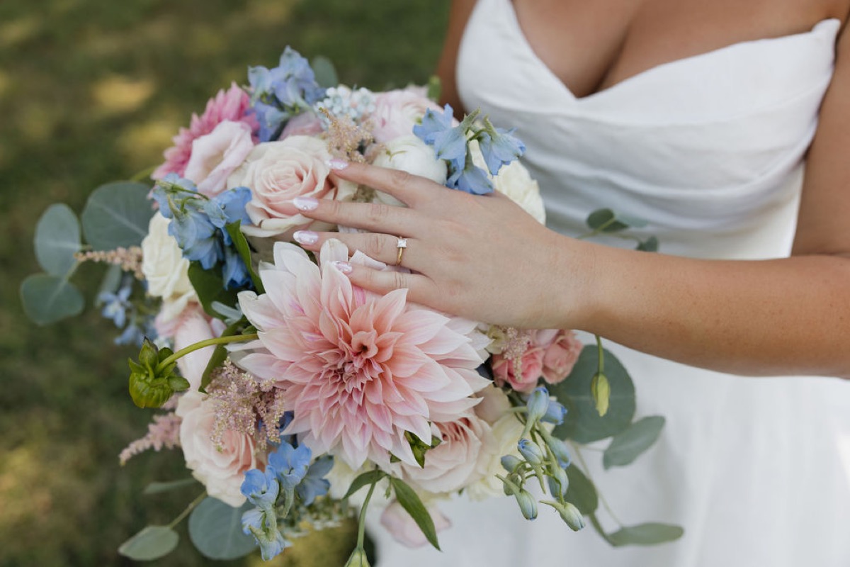 Lilly’s ring hand rests on a pink dahlia and blue bouquet at Lilly & Jackson’s Covey Creek wedding