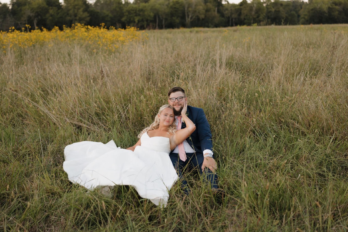Lilly & Jackson sit close in tall grass at Covey Creek, her hand on his face beside yellow wildflowers