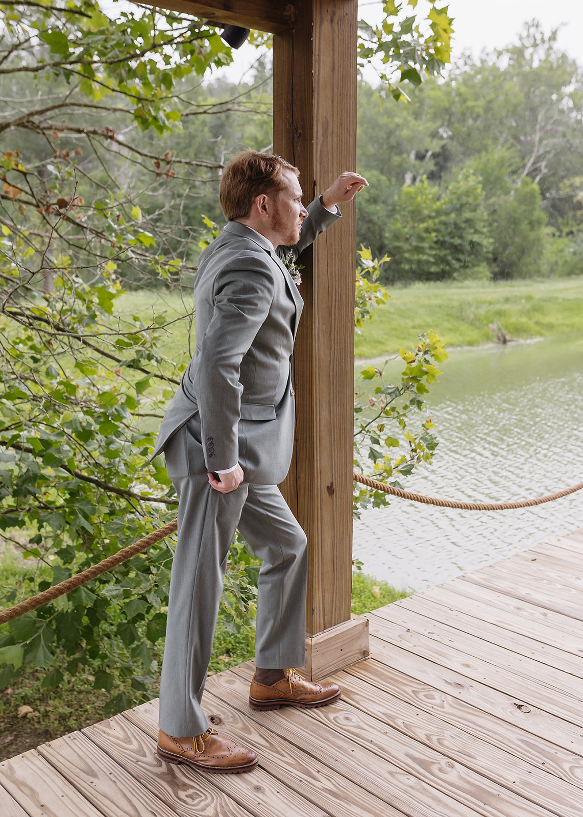 Benjamin leans on a Pond dock post in a grey suit for Meredith & Benjamin's day at Covey Creek