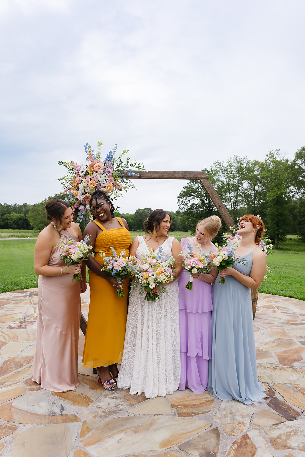 Meredith & Benjamin's bridal party laughs in mixed pastel dresses by the Patio arch at Covey Creek