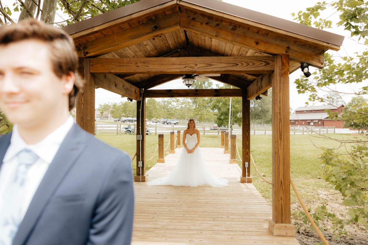 Morgan & Caden first look on Covey Creek’s Pond pavilion dock, bride approaching with red barn beyond