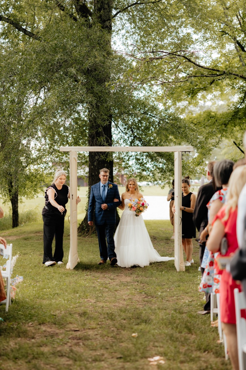 Morgan walks through white freestanding doors at Covey Creek Pond for Morgan & Caden’s ceremony aisle