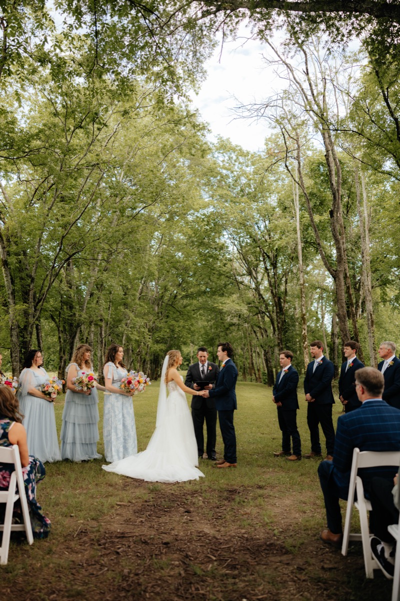 Morgan & Caden exchange vows under Covey Creek’s Tunnel of Love canopy with blue bridesmaids beside them