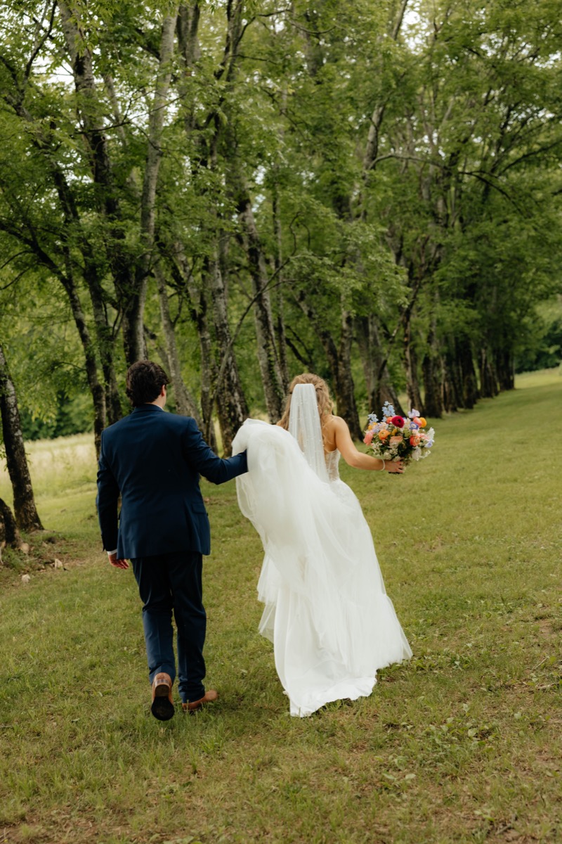 Morgan & Caden walk a grassy field at Covey Creek as he lifts the long train of her white gown